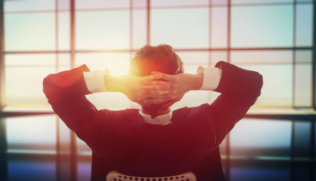 Mann im Anzug lehnt sich entspannt mit verschränkten Händen hinter dem Kopf zurück und blickt durch ein sonniges Bürofenster – Photovoltaik Investment Vorteile